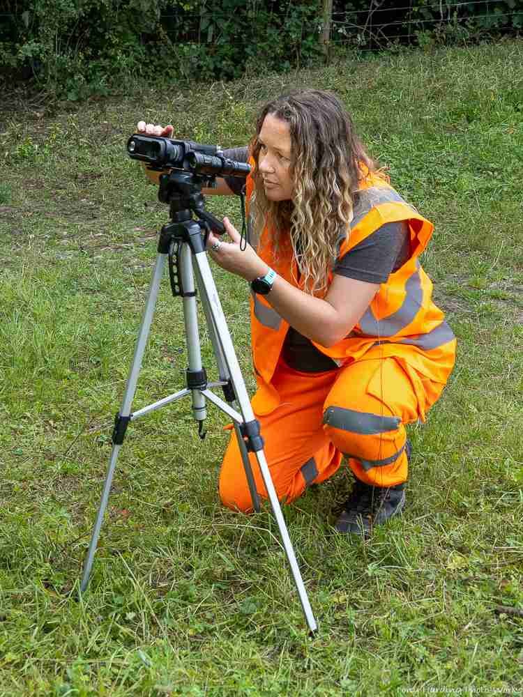 Poppy from PJH Ecology in an orange high visibility outfit looking through a camera on a tripod